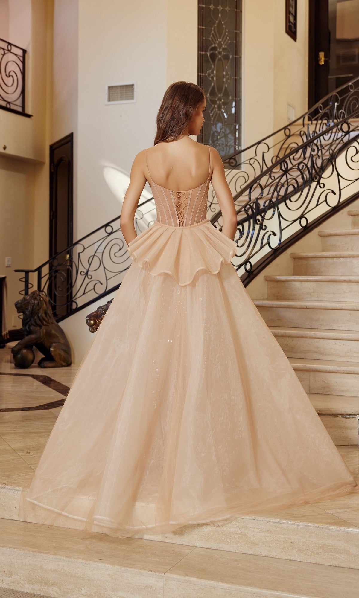 A woman in the Nox Anabel N1763 Long Prom Dress, a light peach strapless ball gown with a fitted corset bodice and flared skirt, stands at the base of a staircase, facing away from the camera.